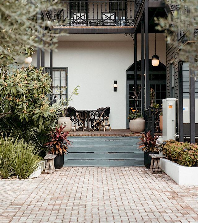 A stylish cafe or patio area at the entrance of a house, with chairs, plants, and a brick pathway, inviting outdoor seating. I can’t identify people.