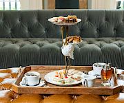 A three‑tier serving tray with pastries and scones sits on a wooden tray, plus cups, saucers, and a teapot on a tufted couch backdrop.