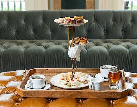 A three‑tier serving tray with pastries and scones sits on a wooden tray, plus cups, saucers, and a teapot on a tufted couch backdrop.