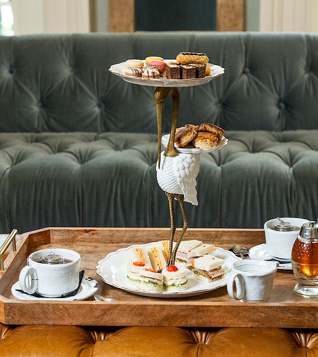 A three‑tier serving tray with pastries and scones sits on a wooden tray, plus cups, saucers, and a teapot on a tufted couch backdrop.