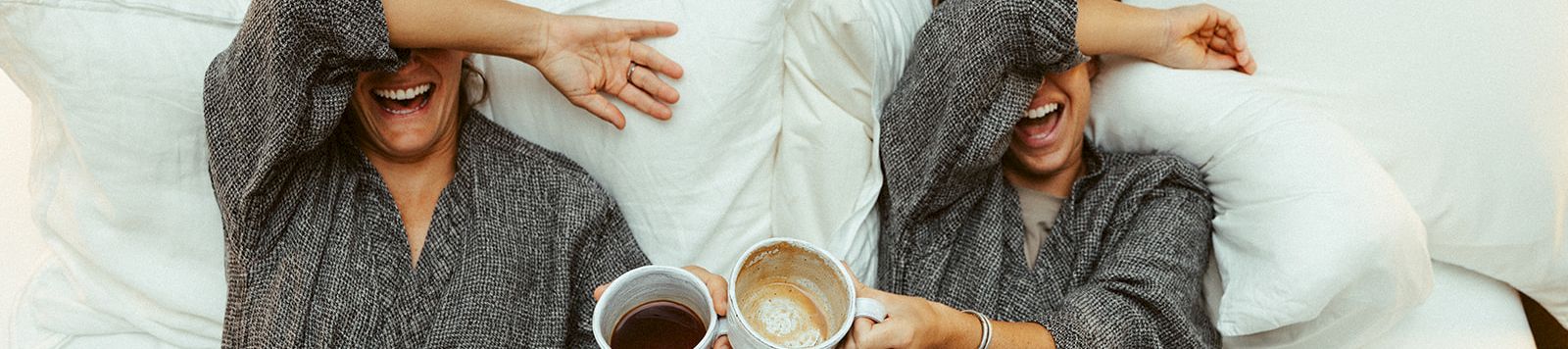 Two women in bathrobes lie in bed, laughing and clinking coffee cups, as they cover their eyes playfully.