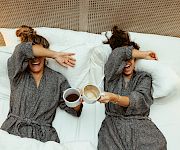 Two women in bathrobes lie in bed, laughing and clinking coffee cups, as they cover their eyes playfully.