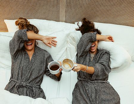 Two women in bathrobes lie in bed, laughing and clinking coffee cups, as they cover their eyes playfully.