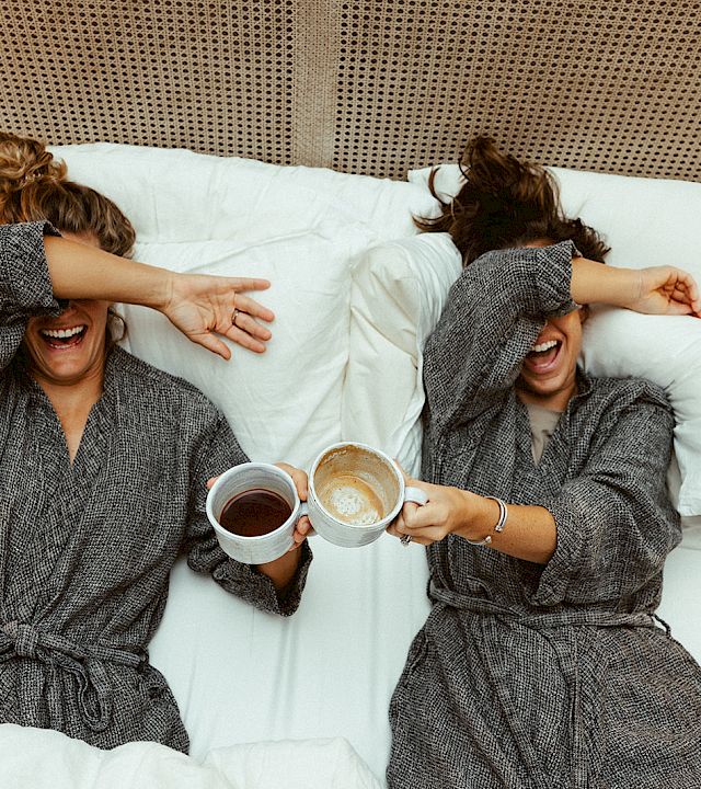 Two women in bathrobes lie in bed, laughing and clinking coffee cups, as they cover their eyes playfully.