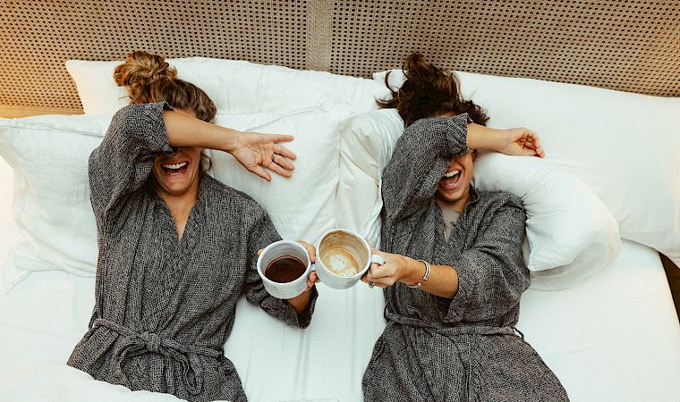 Two women in bathrobes lie in bed, laughing and clinking coffee cups, as they cover their eyes playfully.