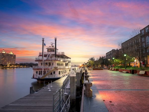 A moored river cruise ship sits by a waterfront promenade at sunset, with city lights glowing along the walkway.