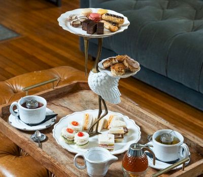 A wooden tray with a three-tiered serving dish filled with pastries and sandwiches, accompanied by tea and creamers.