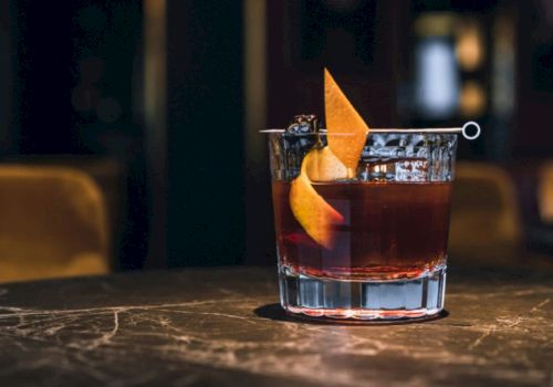 A cocktail in a lowball glass, topped with an orange peel twist, on a bar table.
