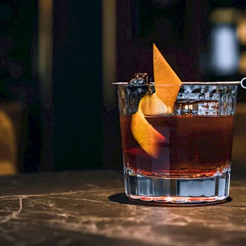 A cocktail in a lowball glass, topped with an orange peel twist, on a bar table.