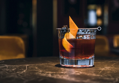 A short whiskey cocktail in a glass with ice and an orange peel garnish, sitting on a bar table.