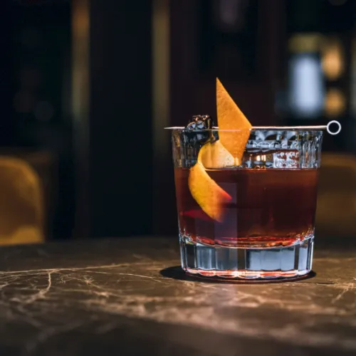 A short whiskey cocktail in a glass with ice and an orange peel garnish, sitting on a bar table.