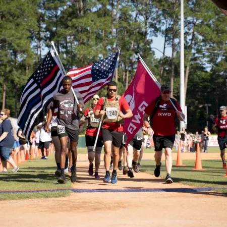 A group of runners sprints on a track as the U.S. flag and a Canadian flag wave behind them.