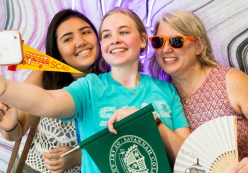 Three smiling women pose for a selfie at an event, with fans and a green Savannah folder in hand.