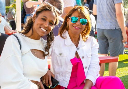 Two women smiling outdoors at a sunny event; one wears blue mirrored sunglasses, both in white outfits with a bright pink bow.