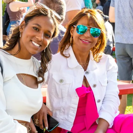 Two women sit outdoors smiling in white outfits, one wearing teal sunglasses and bright pink bottoms at a sunny event.