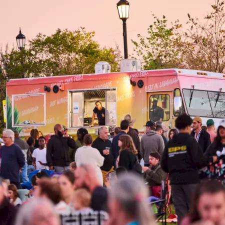 A crowd gathers at sunset near a large pink bus, where people stand and move around in the street.