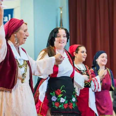 A group of women in colorful traditional folk outfits dance onstage, with one performer raising her arm in celebration.