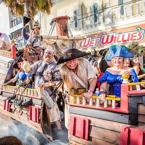 A pirate-themed parade boat with excited costumed performers on board, cheering in front of a &ldquo;Wild Wild&rdquo; float, in sunny street view.