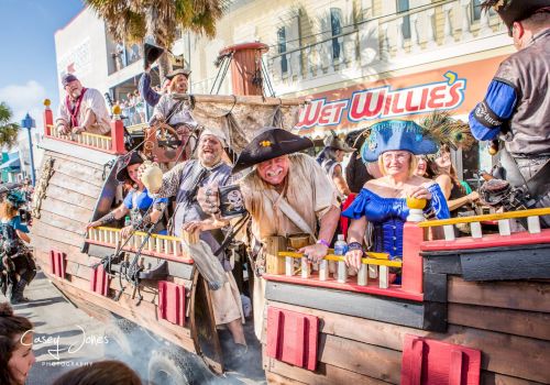 A pirate-themed parade boat with excited costumed performers on board, cheering in front of a &ldquo;Wild Wild&rdquo; float, in sunny street view.