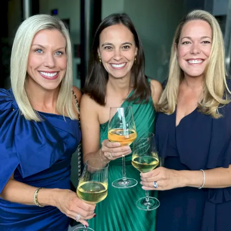 Three smiling women in blue/green dresses toast with white wine glasses at an event.