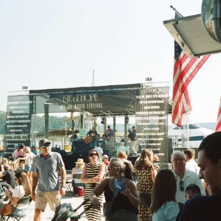 Crowd gathers at an outdoor music festival with a stage labeled &ldquo;Isle of Hope&rdquo; and American flags waving nearby.