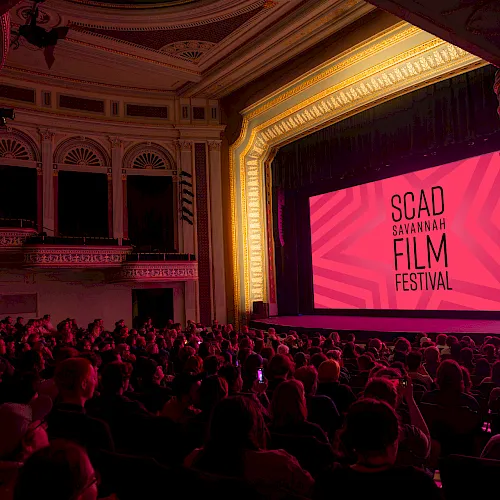 A packed theater auditorium with a big screen reading &ldquo;SGAD Savannah Film Festival,&rdquo; lit in pink/red tones, during a film event.