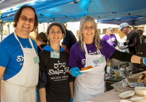 Three people in aprons smile at an outdoor kosher food stall, serving dishes under a blue tent, with other attendees behind them.