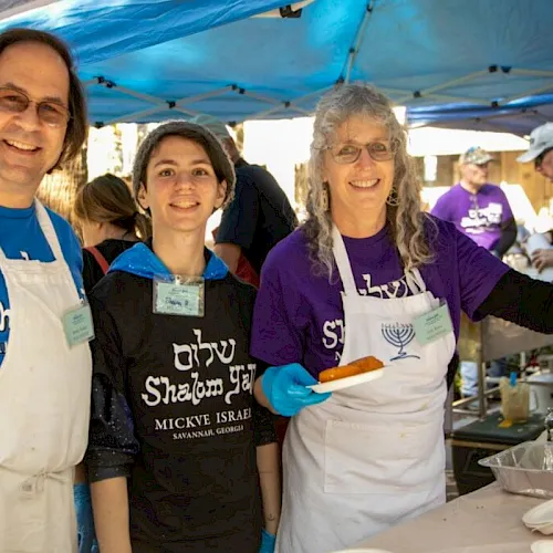 Three people in aprons smile at an outdoor kosher food stall, serving dishes under a blue tent, with other attendees behind them.
