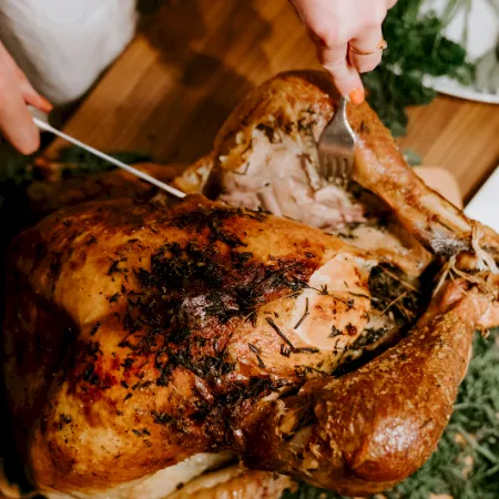 A person carves a roasted turkey on a wooden cutting board with herbs nearby, using carving forks and a knife.