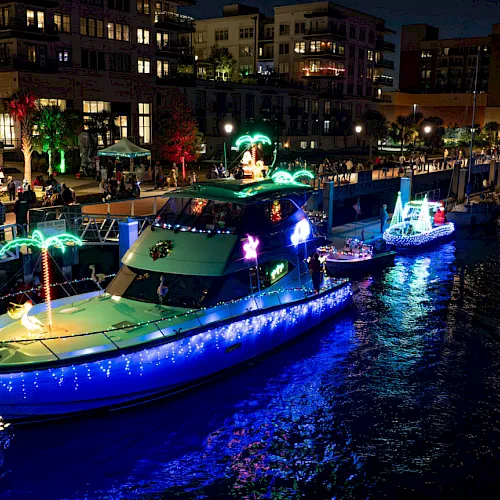 At night, a lighted holiday parade of boats floats along a canal beside lit waterfront buildings.