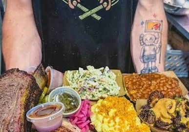 A man in a &ldquo;SlowFire BBQ&rdquo; shirt serves a spread of smoked meats, sides, and sauces, including ribs, brisket, chicken, and pulled pork.
