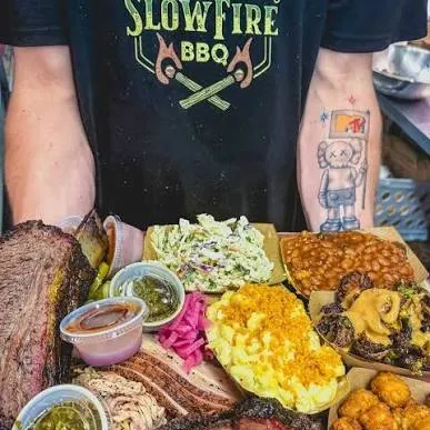 A man in a &ldquo;SlowFire BBQ&rdquo; shirt serves a spread of smoked meats, sides, and sauces, including ribs, brisket, chicken, and pulled pork.
