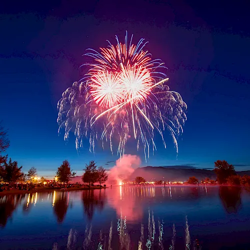 A bright multicolored firework explodes above a calm lake, reflecting in the water as trees and night sky frame the scene.