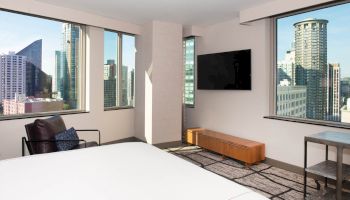 A modern hotel room features a large bed, city views through windows, a wall-mounted TV, an armchair, and a small table.