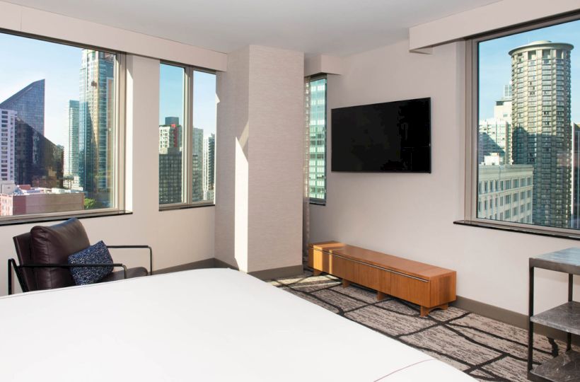 A modern hotel room features a large bed, city views through windows, a wall-mounted TV, an armchair, and a small table.