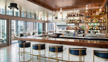 A modern, upscale bar with blue stools, a well-stocked liquor shelf, pendant lighting, and large windows overlooking a cityscape on a sunny day.