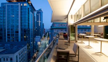 An outdoor terrace overlooking a city street with modern high-rise buildings at dusk. The terrace has tables and chairs arranged for dining.