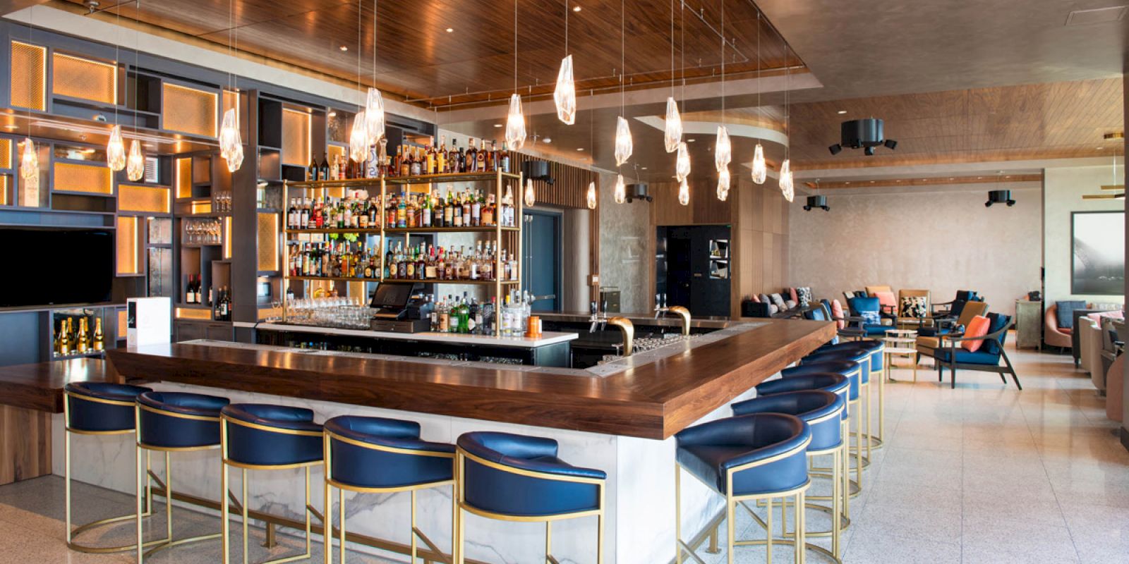 This image shows a modern bar with a sleek design, featuring a long wooden counter, bar stools, hanging lights, and shelves filled with bottles.
