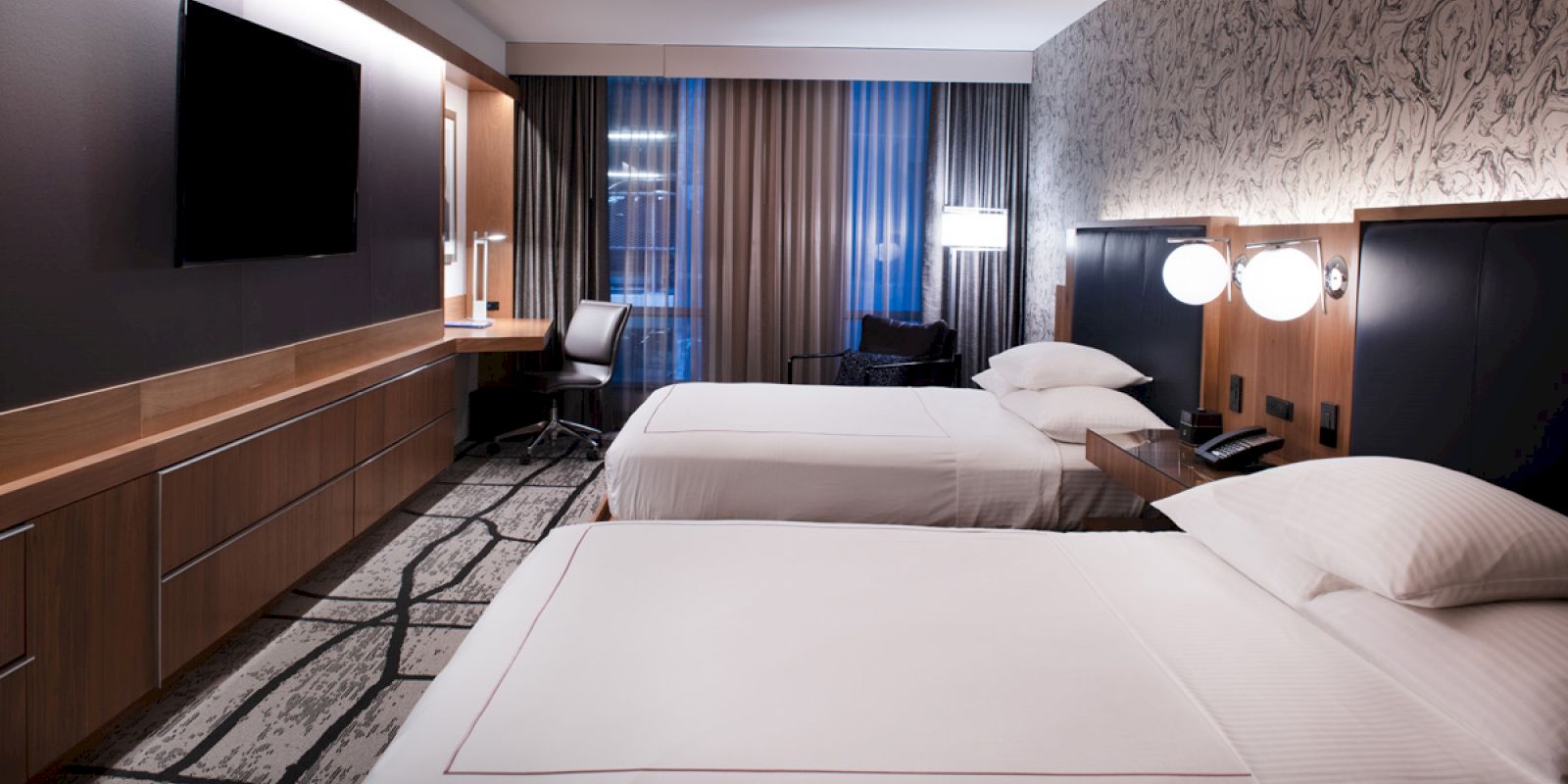 A modern hotel room with two beds, a wall-mounted TV, a desk with a chair, and bedside lamps, all in contemporary decor.