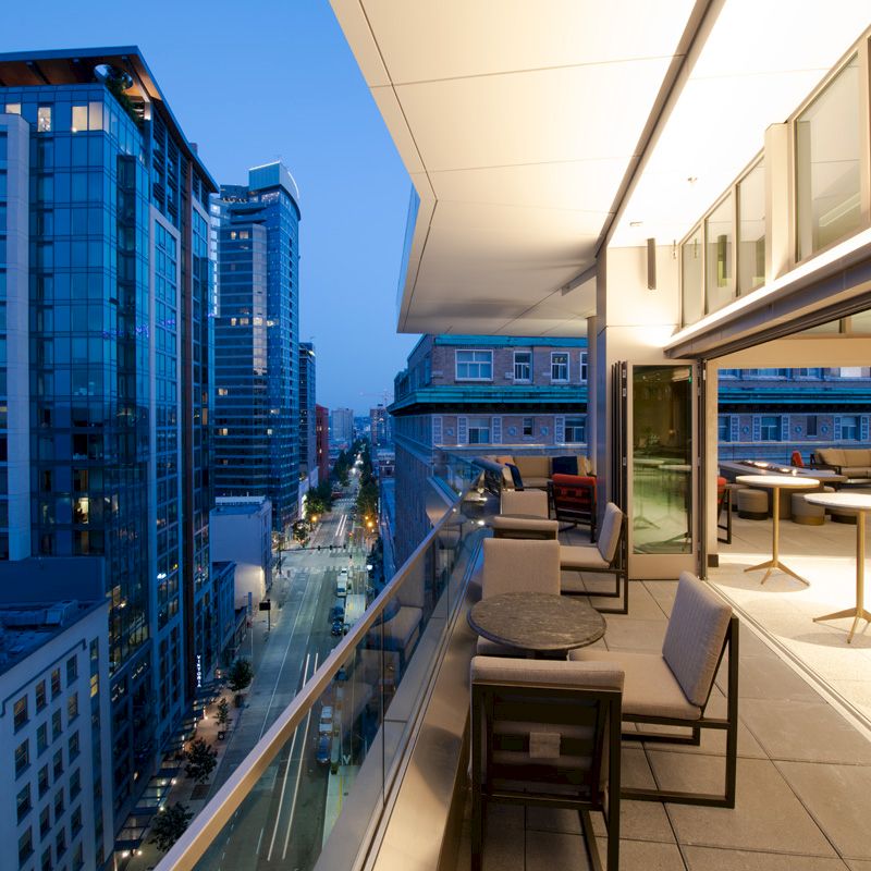 An outdoor urban patio at dusk with modern furnishings overlooks city buildings and streets under a blue sky, featuring a high-rise view.