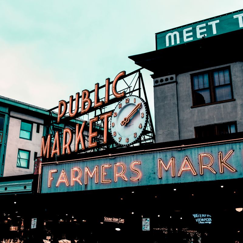 The image shows the iconic neon sign of a public market and farmers market in an urban area, with buildings in the background and a clock in the center.