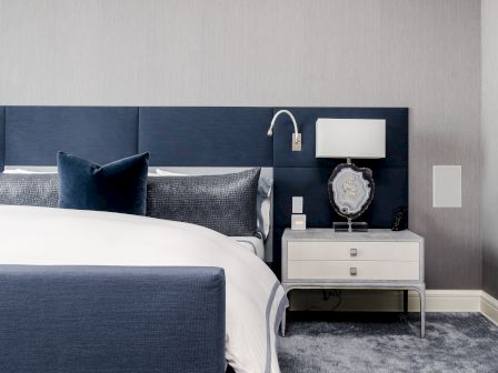 A modern bedroom featuring a bed with a blue headboard and white bedding. There's a nightstand with a lamp and a decorative item.