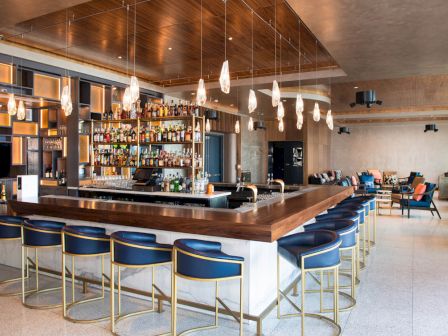 The image shows a modern bar with a wooden counter, blue chairs, and hanging lights. The shelves behind the bar are stocked with various bottles.
