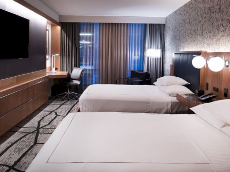 A modern hotel room with two beds, a wall-mounted TV, a work desk, and contemporary decor with muted color tones and soft lighting.