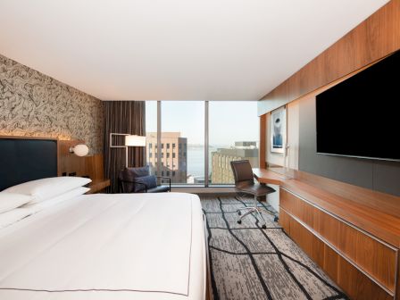 A modern hotel room with a large bed, desk, chair, wall-mounted TV, and a window offering a city view with buildings and partially a body of water.