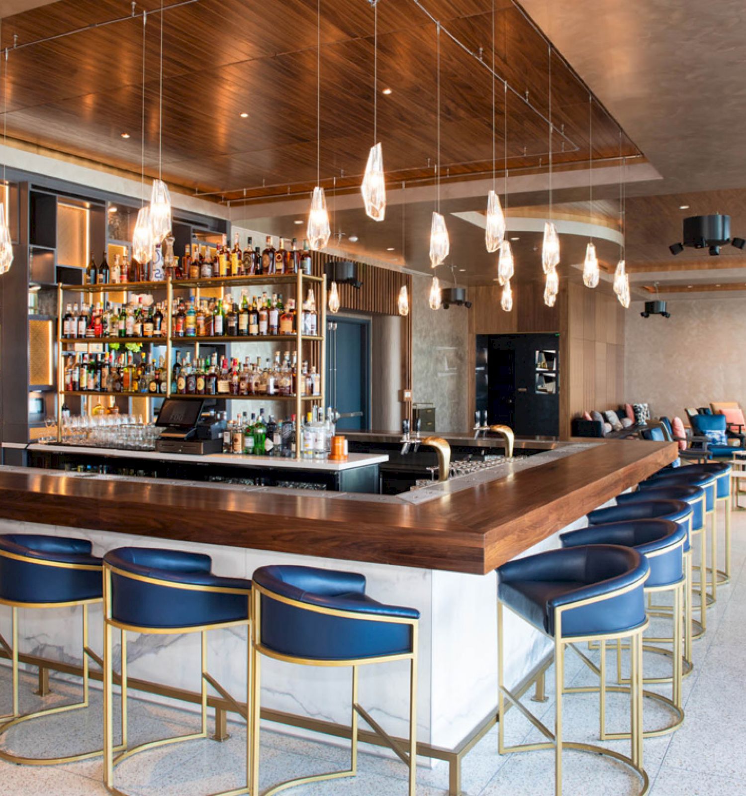 A modern bar with a wooden top counter, blue cushioned stools, hanging lights, and a well-stocked shelves for bottles in the background.