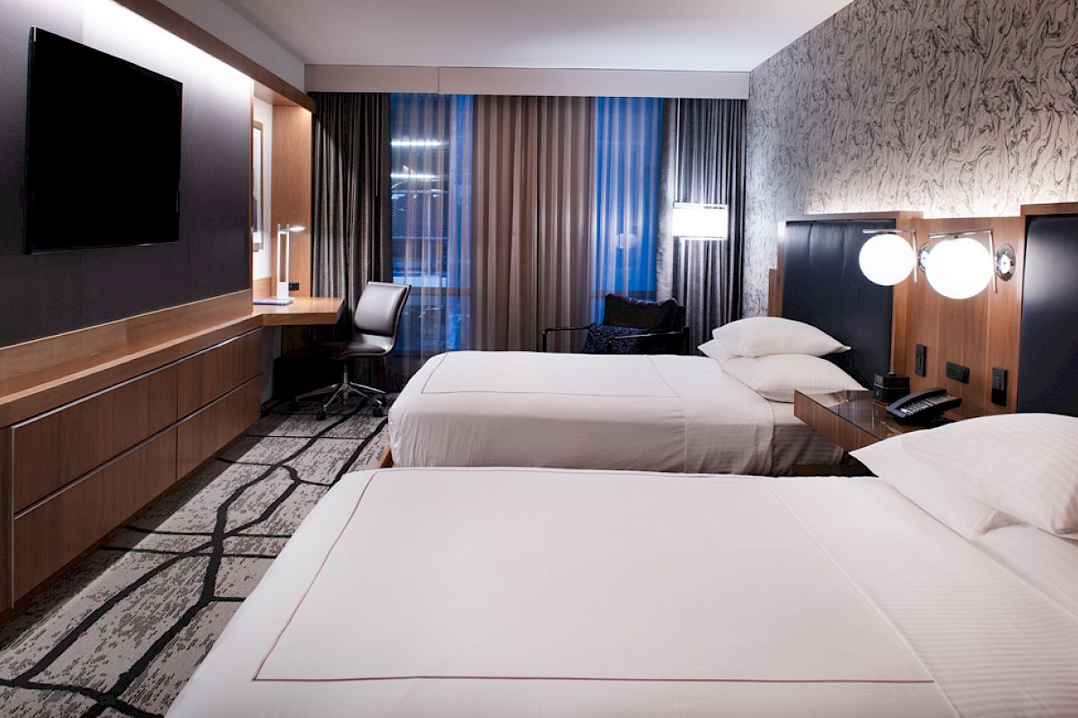 A modern hotel room with two double beds, a large TV, a desk with a chair, and contemporary lighting and decor.
