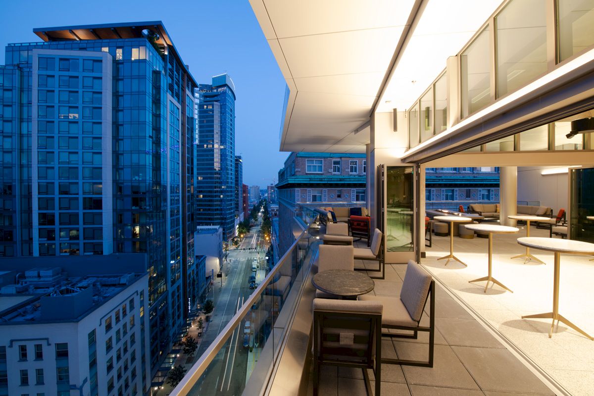 A modern, stylish balcony with seating overlooks a cityscape of tall buildings at dusk, featuring a blend of indoor and outdoor spaces.