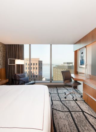 A modern hotel room with a large bed, wall-mounted TV, desk and chair, armchair, and a window offering a cityscape view in the background.