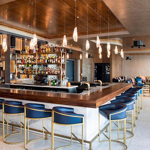This image shows a modern bar with blue chairs, a fully stocked selection of drinks, overhead pendant lighting, and a lounge area in the background.